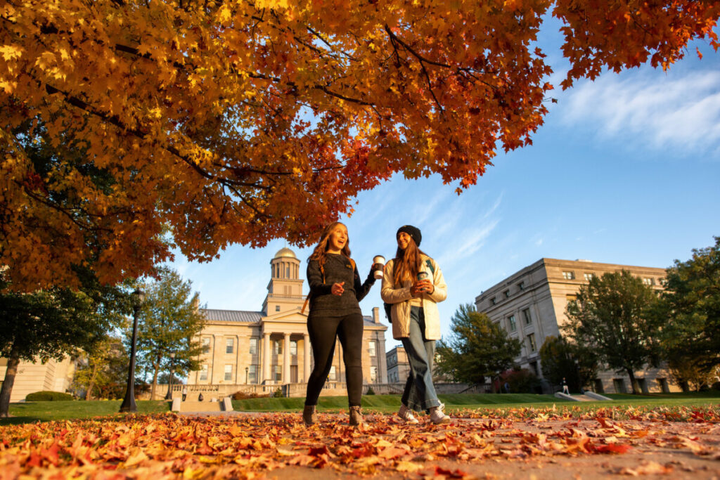 Two students walking in front of the Old Capitol building on a crisp fall day. The leaves in the foreground are brightly colored and have begun to fall onto the sidewalk where students are walking.