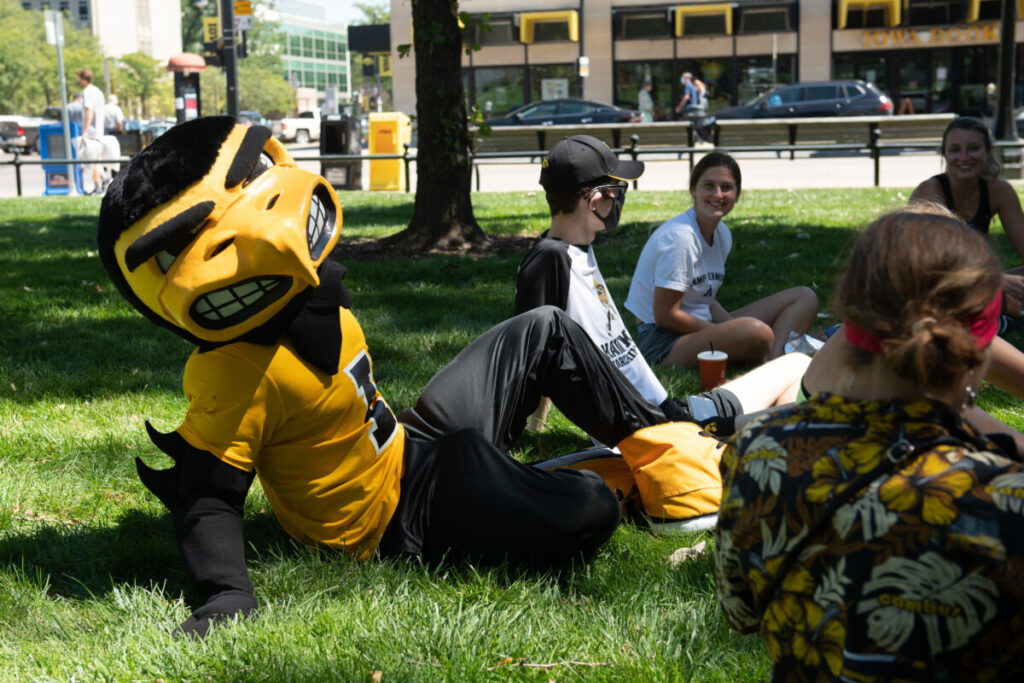 Herky (Iowa's mascot) sitting on grass with a group of students in the shade.