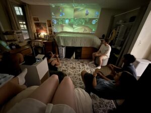 Displayed on the wall is a projected video of Mario kart, surrounded by different people sitting on the floor, bed, and chairs. It's dark, only illuminated by the open door and projection. 