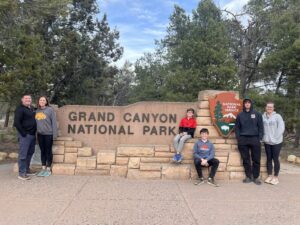 Kiah stands to the viewer's right of a sign reading "Grand Canyon National Park" with her brothers. Her parents stand to the left. 