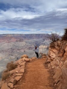 Kiah stands on the edge of a trail leading down into the grand canyon. She is far from the camera, looking at it, while the canyon drops away for miles behind her. 