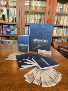 White bookmarks and blue books fan across a wooden table. The original painting that is the book cover, a whale with a gradient blue background, is also propped upright on the table. 