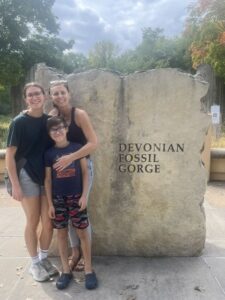 Kiah, her mother, and her little brother Logan stand to the right of a rock, smiling at the camera. The rock reads "Devonian Fossil Gorge."